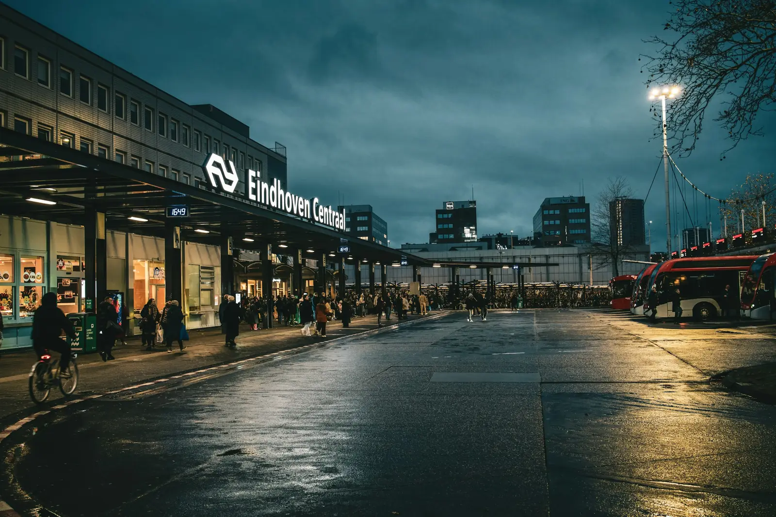 Stationsplein van Eindhoven Centraal in de schemering, met reizigers, fietsers en bussen op natte straat